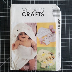 May include: A McCall's Crafts pattern package featuring a baby wrapped in a white hooded towel with a bear face design. The package also shows examples of baby accessories like hats, mittens, and booties. The number 3697 is visible.