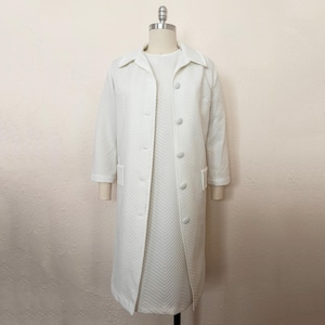 May include: A vintage white coat and dress set. The coat is long, with a collared neckline, button closures, and pockets. The dress underneath has a similar design. Both pieces feature a textured, chevron pattern. The set is displayed on a mannequin.