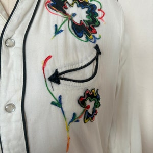 Detail view of 1960s vintage white rayon pearl snap western shirt  with navy trim and multicolored embroidery by H bar C, size small to medium. Showing the curved chest pocket and multicolor embroidery with some breaks in the thread.