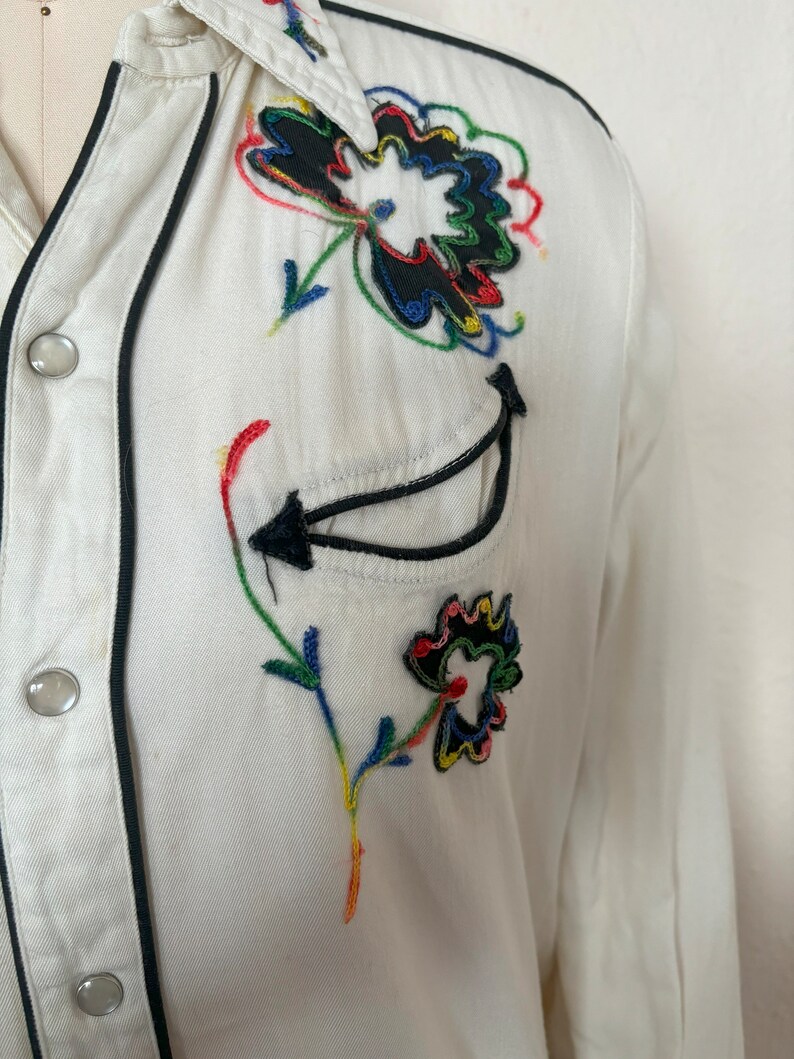 Detail view of 1960s vintage white rayon pearl snap western shirt  with navy trim and multicolored embroidery by H bar C, size small to medium. Showing the curved chest pocket and multicolor embroidery with some breaks in the thread.