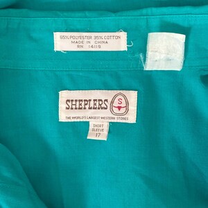 Interior detail view of 1980s vintage teal pearl snap short sleeve western shirt, by Sheplers, size medium to large. Showing the Sheplers label, fabric composition and size 17 tag.