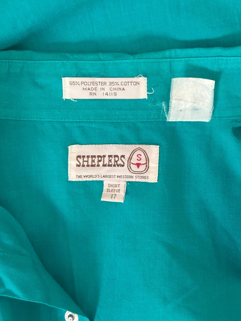 Interior detail view of 1980s vintage teal pearl snap short sleeve western shirt, by Sheplers, size medium to large. Showing the Sheplers label, fabric composition and size 17 tag.