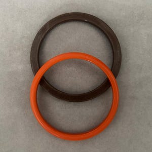 May include: Two circular rubber gaskets. One is a dark brown color, and the other is bright orange. The gaskets are positioned on top of each other, with the orange one in front. The background is a light gray color.