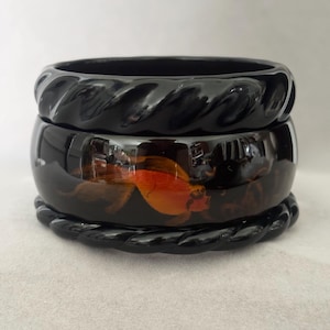 Puede incluir: Tres pulseras negras apiladas. Las pulseras superior e inferior tienen un diseño de cuerda trenzada. La pulsera del medio tiene un fondo oscuro con detalles florales en naranja y marrón.