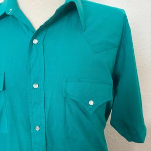 Detail view of 1980s vintage teal pearl snap short sleeve western shirt, by Sheplers, size medium to large. Showing the pointed yoke, and pockets as well as the pearl snaps.