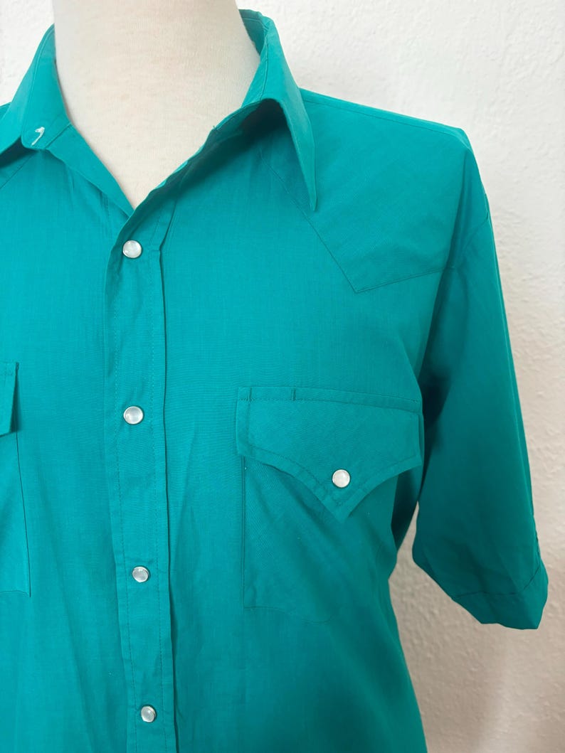 Detail view of 1980s vintage teal pearl snap short sleeve western shirt, by Sheplers, size medium to large. Showing the pointed yoke, and pockets as well as the pearl snaps.