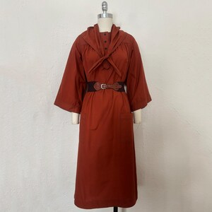 May include: A rust-colored dress with a unique collar and a brown belt. The dress features a button-down front, long sleeves, and pockets. The belt has a buckle closure. The dress is displayed on a mannequin.