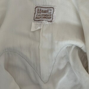 Interior detail view of 1960s vintage white rayon pearl snap western shirt  with navy trim and multicolored embroidery by H bar C, size small to medium. Showing the curved chest pocket and multicolor embroidery with some breaks in the thread. Showing the H Bar C California Ranchwear label.