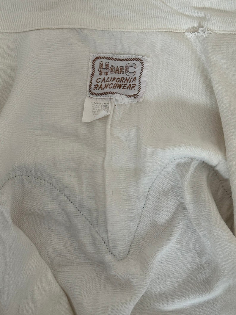 Interior detail view of 1960s vintage white rayon pearl snap western shirt  with navy trim and multicolored embroidery by H bar C, size small to medium. Showing the curved chest pocket and multicolor embroidery with some breaks in the thread. Showing the H Bar C California Ranchwear label.