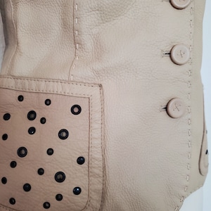 Detail view of 1990s topstitched metal eyelet and stud embellished buff leather blazer by Donald Pliner, extra small to small, showing the topstitching, self covered leather buttons, and patch pockets embellished with black eyelets and studs.