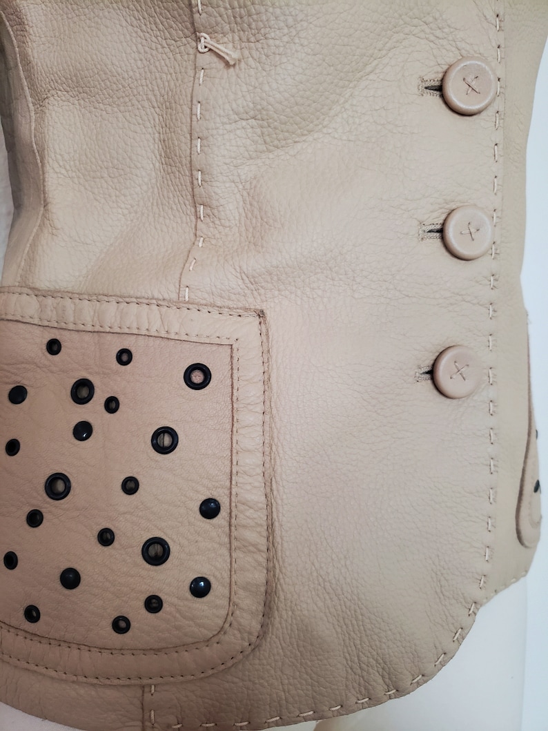 Detail view of 1990s topstitched metal eyelet and stud embellished buff leather blazer by Donald Pliner, extra small to small, showing the topstitching, self covered leather buttons, and patch pockets embellished with black eyelets and studs.