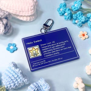 Könnte beinhalten: Ein blauer Acryl-Schlüsselanhänger mit dem Text "Holy Symbol" und Spieldetails. Der Schlüsselanhänger hat einen silbernen Verschluss und ist von gehäkelten Blumen in Blau-, Weiß- und Gelbtönen umgeben. Ein Spielzubehör.