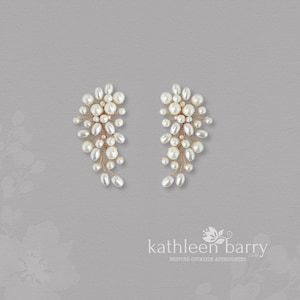 Peut inclure: Une paire de boucles d'oreilles en perles. Chaque boucle d'oreille présente un groupe de perles blanches de tailles variées, disposées sur un fil doré délicat. Les boucles d'oreilles ont un design en cascade, les perles étant plus clairsemées vers le bas. Le fond est gris clair.