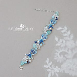 Puede incluir: Un delicado brazalete de plata con cuentas de vidrio azul y blanco, conchas marinas y estrellas de mar. El brazalete está adornado con pequeñas flores blancas y cristales azules. El brazalete es una pieza de joyería hermosa y única que sería perfecta para una boda de verano o unas vacaciones en la playa.