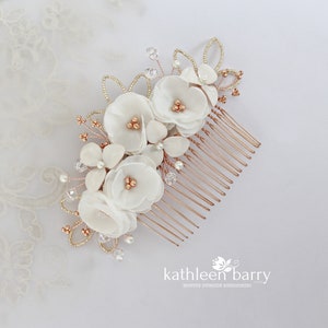 May include: White floral hair comb with rose gold accents. The comb features white fabric flowers, pearl and crystal embellishments, and a rose gold metal comb.