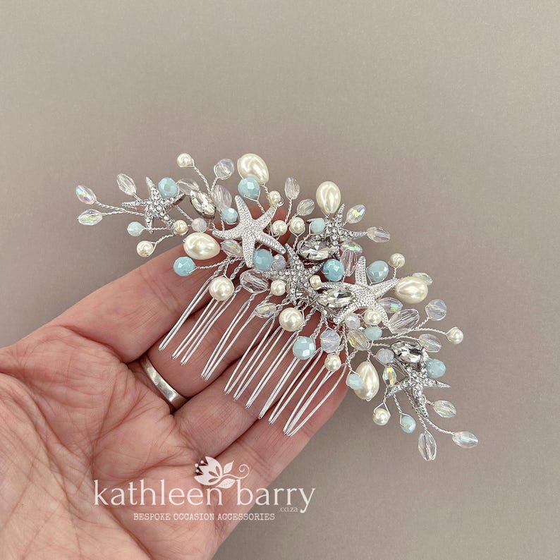 May include: A silver hair comb featuring a coastal design with multiple starfish accents, light blue beads, clear crystals, and white faux pearls arranged on thin silver wire branches. The text kathleen barry is printed at the bottom.