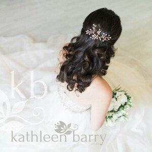 May include: A bride with long, dark curly hair wears a white strapless wedding dress and a gold hair comb with small white pearls. She holds a bouquet of white flowers.