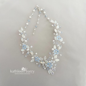 Op de afbeelding: Een zilverkleurige draadketting met lichtblauwe bloemaccenten, witte parels en heldere kristallen. Het ontwerp bevat metalen bladvormen en kleine blauwe kralen die in een rankachtig patroon zijn gerangschikt. De tekst kathleen barry is zichtbaar.