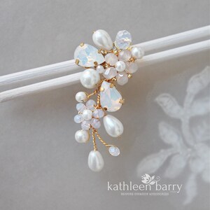 May include: A bridal hair accessory featuring a cluster of white pearls, opalescent teardrop-shaped crystals, and small pink beads. The design is accented with gold wire and is mounted on a white bar. The accessory is for special occasions.