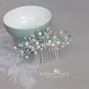 May include: A silver hair comb with a delicate design featuring green and white beads. The comb is set against a light green background.