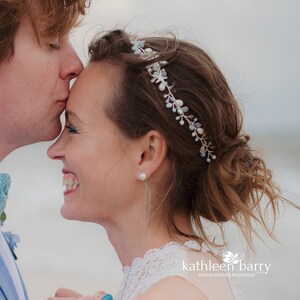 May include: A bride smiles as the groom kisses her forehead. She is wearing a white lace dress and a delicate silver and blue hairpiece. The groom is wearing a blue suit and a blue bow tie.