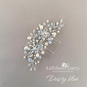 May include: A silver hair comb featuring a cluster of starfish charms, white pearls, and light blue crystals on thin wire branches. The text "kathleen barry BESPOKE OCCASION ACCESSORIES" and "Dusty blue" is printed in white in the bottom right corner.