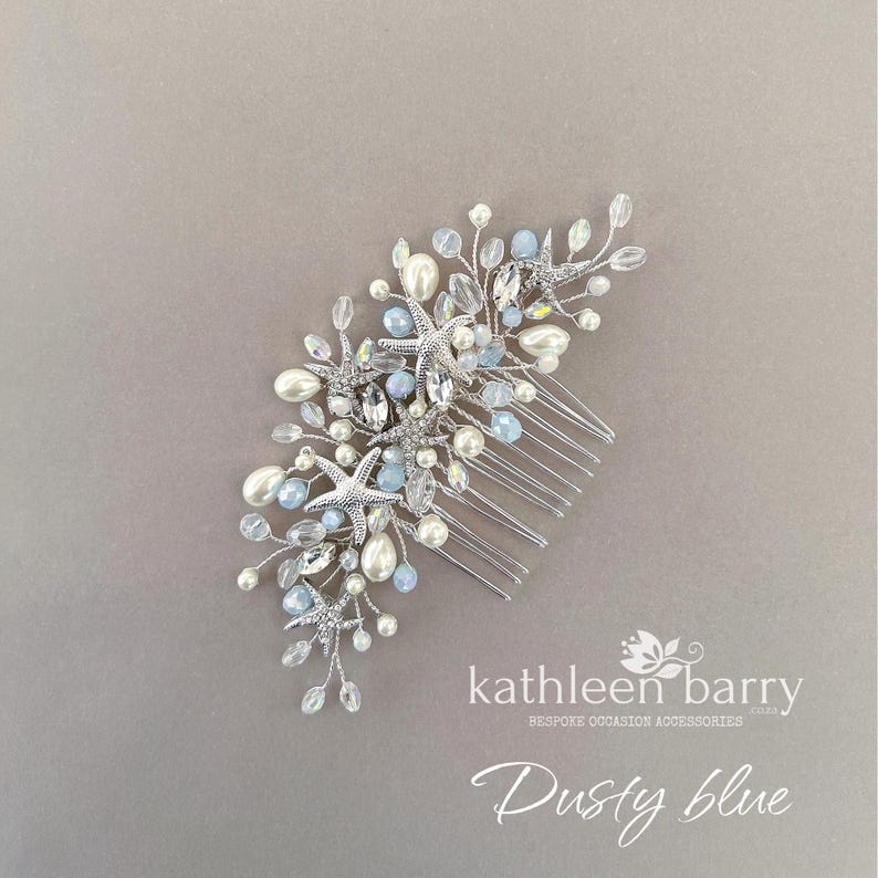 May include: A silver hair comb featuring a cluster of starfish charms, white pearls, and light blue crystals on thin wire branches. The text "kathleen barry BESPOKE OCCASION ACCESSORIES" and "Dusty blue" is printed in white in the bottom right corner.