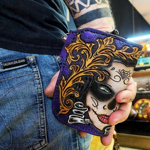 Könnte beinhalten: Eine violette Ledergeldbörse mit einer detaillierten Schnitzerei eines Frauen Gesichts mit einem Zucker Schädel Design. Die Schnitzerei ist in Gold und Schwarz, und die Frau trägt eine Maske. Die Geldbörse hat ein florales Design an den Rändern.