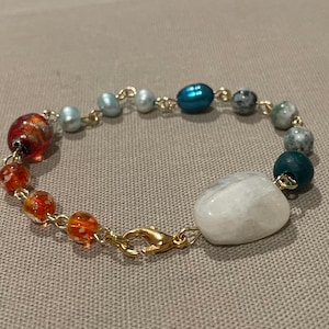 Op de afbeelding: Een kralenarmband met kleurrijke kralen, waaronder oranje, rode, blauwe en groene kralen. De armband heeft een goudkleurige ketting en sluiting, en een grote, witte, afgeronde steen als middelpunt.