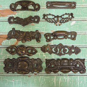 May include: A collection of antique brass drawer pulls and handles in various ornate designs. The hardware features intricate scrollwork, floral motifs, and keyhole details. The pulls are arranged on a distressed, weathered, light green wooden surface.