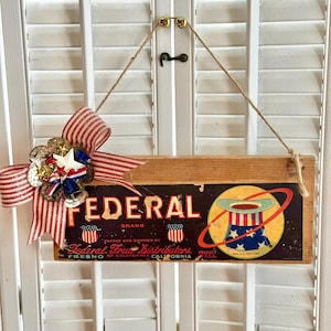 May include: Wooden sign with the word "FEDERAL" in bold letters, featuring a vintage design with patriotic imagery. A red, white, and blue bow and star embellishment adorns the top left corner. The sign is suspended by a jute rope.
