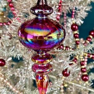 May include: A teardrop-shaped glass ornament featuring a vibrant iridescent finish with deep purple, magenta, and golden hues. The ornament has a rounded central body, a tapered finial at the bottom, and a decorative neck with a small loop at the top.