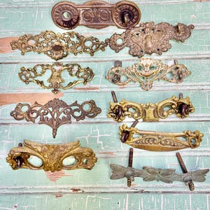 May include: A collection of antique brass drawer pulls and handles in various ornate designs. The hardware features intricate scrollwork, floral motifs, and aged patinas. The pulls are mounted on a distressed, light green wooden surface.