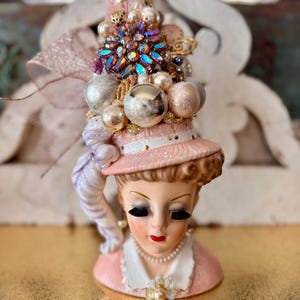 May include: A vintage-style ceramic figurine of a woman wearing a pink hat adorned with silver and pink ornaments, a large jeweled brooch, and pearls. The figurine has closed eyes, red lips, and a pearl necklace. The background is a gold surface.