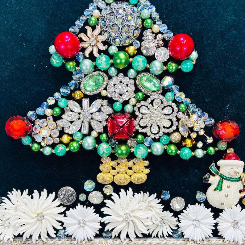 May include: A Christmas tree crafted from green, red and white beads, rhinestones and jewellery. The tree is adorned with a snowman ornament and white flowers at the base.