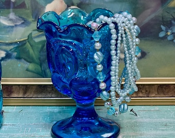 May include: A vibrant cobalt blue glass pedestal dish with a scalloped edge, adorned with star and moon patterns. It displays light blue beaded necklaces. The background features a painting with floral motifs.