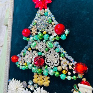 May include: A Christmas tree crafted from a variety of jewellery and trinkets, including rhinestones, beads, and bells. The tree is adorned with red and green baubles and is set against a dark green backdrop.