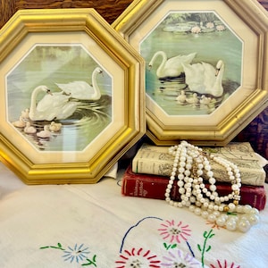 May include: Two framed swan prints with gold octagonal frames. The prints depict white swans and their cygnets on a lake. A pearl necklace and embroidered floral tablecloth are also visible.