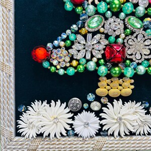 May include: A framed Christmas tree made of various colourful jewels, rhinestones, and beads. The tree is decorated with green, red, and gold accents. The bottom of the frame is decorated with white flowers.