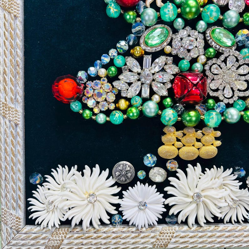 May include: A framed Christmas tree made of various colourful jewels, rhinestones, and beads. The tree is decorated with green, red, and gold accents. The bottom of the frame is decorated with white flowers.