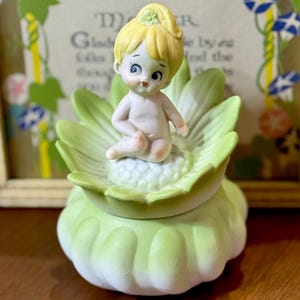 May include: A vintage porcelain figurine of a seated cherub with yellow hair, atop a light green flower-shaped container. The cherub is nude, with delicate facial features. The container has a matching lid and is set against a blurred background.