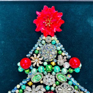 May include: A Christmas tree crafted from an assortment of colourful jewels, beads, and rhinestones. The tree is adorned with a red poinsettia at the top.