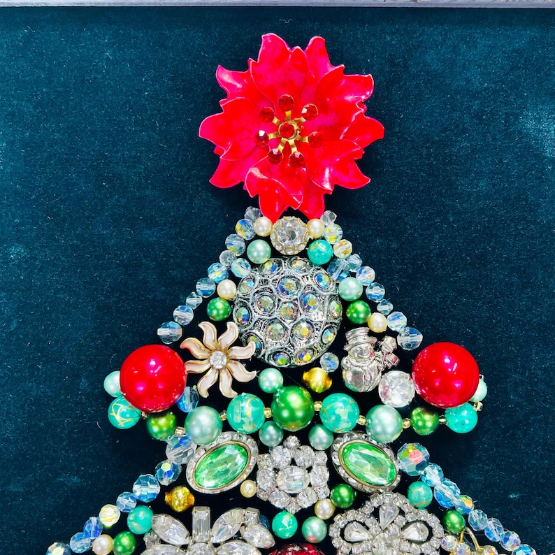 May include: A Christmas tree crafted from an assortment of colourful jewels, beads, and rhinestones. The tree is adorned with a red poinsettia at the top.