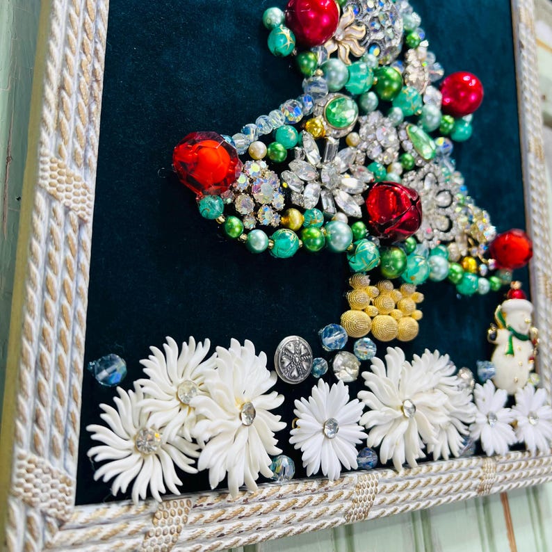 May include: A framed Christmas tree made of rhinestones, beads, and other decorative elements. The tree is on a dark green background and is surrounded by white flowers.