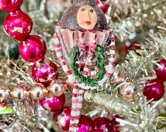 May include: A Christmas tree ornament shaped like a cupcake with a human face, dark brown hair, and red lips. The cupcake base is pink with white stripes, and the figure has long, thin, red and white striped legs and arms holding a small green wreath.