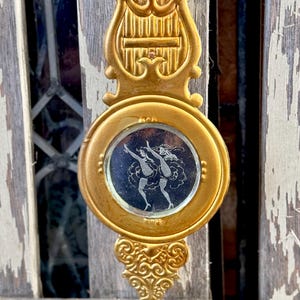 May include: A gold-colored metal ornament with a round mirror featuring a black and white image of two dancers. The ornament has a decorative top shaped like a lyre.