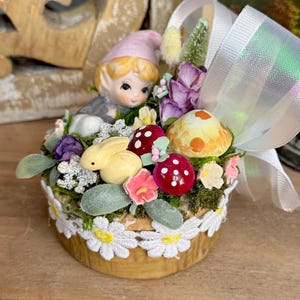 May include: A small ceramic gnome figurine with a pink hat sits in a miniature flower pot with a white lace trim. The pot is filled with artificial flowers, moss, and a small yellow bunny figurine. A white satin ribbon is tied around the pot.