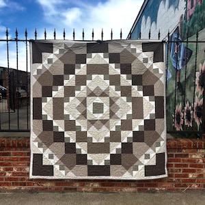 Puede incluir: Un edredón cuadrado con un patrón geométrico en tonos marrones, beige y blanco cuelga de una valla de metal negro. El diseño del edredón presenta cuadrados, rectángulos y triángulos dispuestos simétricamente. El fondo incluye una pared de ladrillo y un mural.