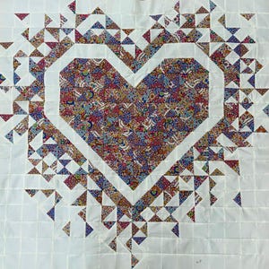 May include: A handmade quilt featuring a large heart design. The heart is filled with a colorful floral pattern, surrounded by a white border and white triangles. The background is white, with a grid pattern.