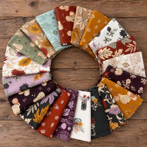 May include: A circular arrangement of twenty folded fabric squares featuring various autumn-themed patterns including sunflowers, pumpkins, leaves, and floral designs in shades of orange, brown, green, purple, black, and white on a wooden surface.